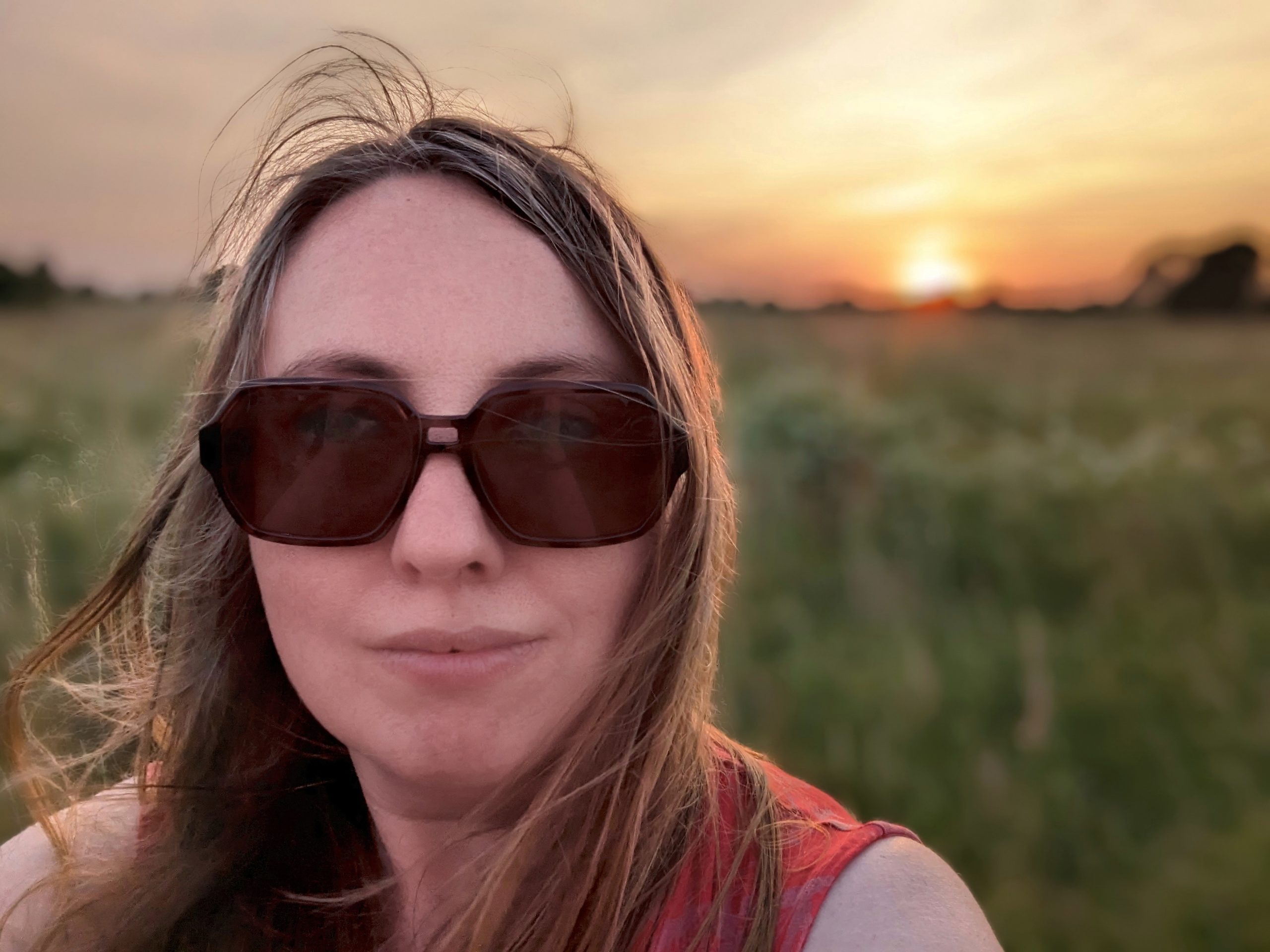 Selfie - woman in sunglasses with sun setting over field in background